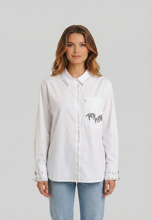 Joven con cabello ondulado que lleva una camisa blanca abotonada con el texto "Tokyo Rocks" en el bolsillo del pecho y vaqueros azul claro.