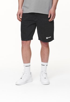 Czarne dżinsowe szorty o luźnym kroju, z naszywką logo na dolnej części nogawki. Nosi się je z białymi butami sportowymi i białymi skarpetkami z logo.