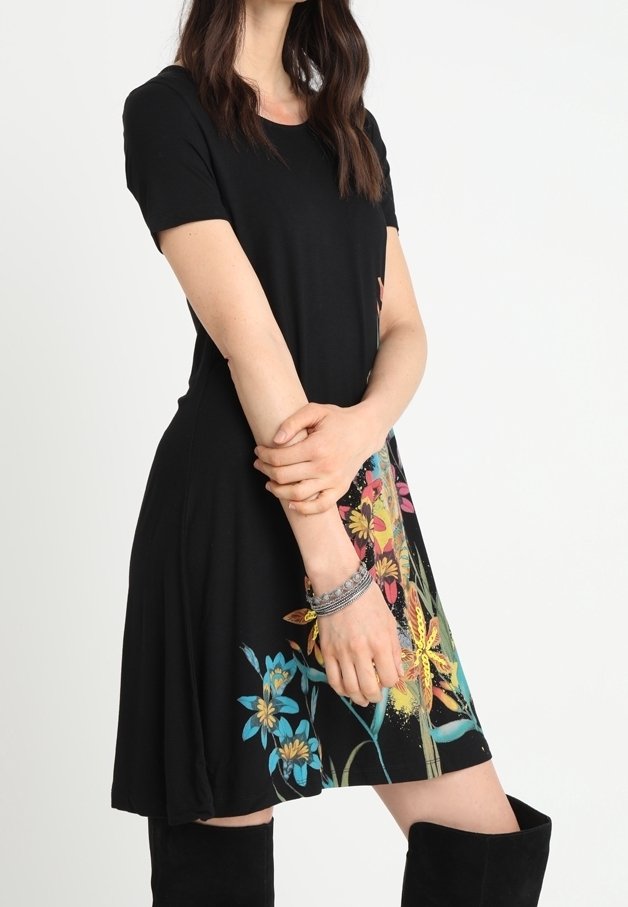 Femme portant une robe noire à manches courtes avec des motifs floraux colorés sur la jupe, associée à des bottes noires montantes jusqu'aux genoux.
