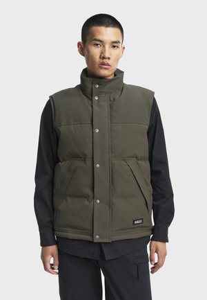 Oliven grøn polstret vest med høj krave, trykknapper, to frontlommer og et logo mærke ved kanten, båret over en sort langærmet skjorte.