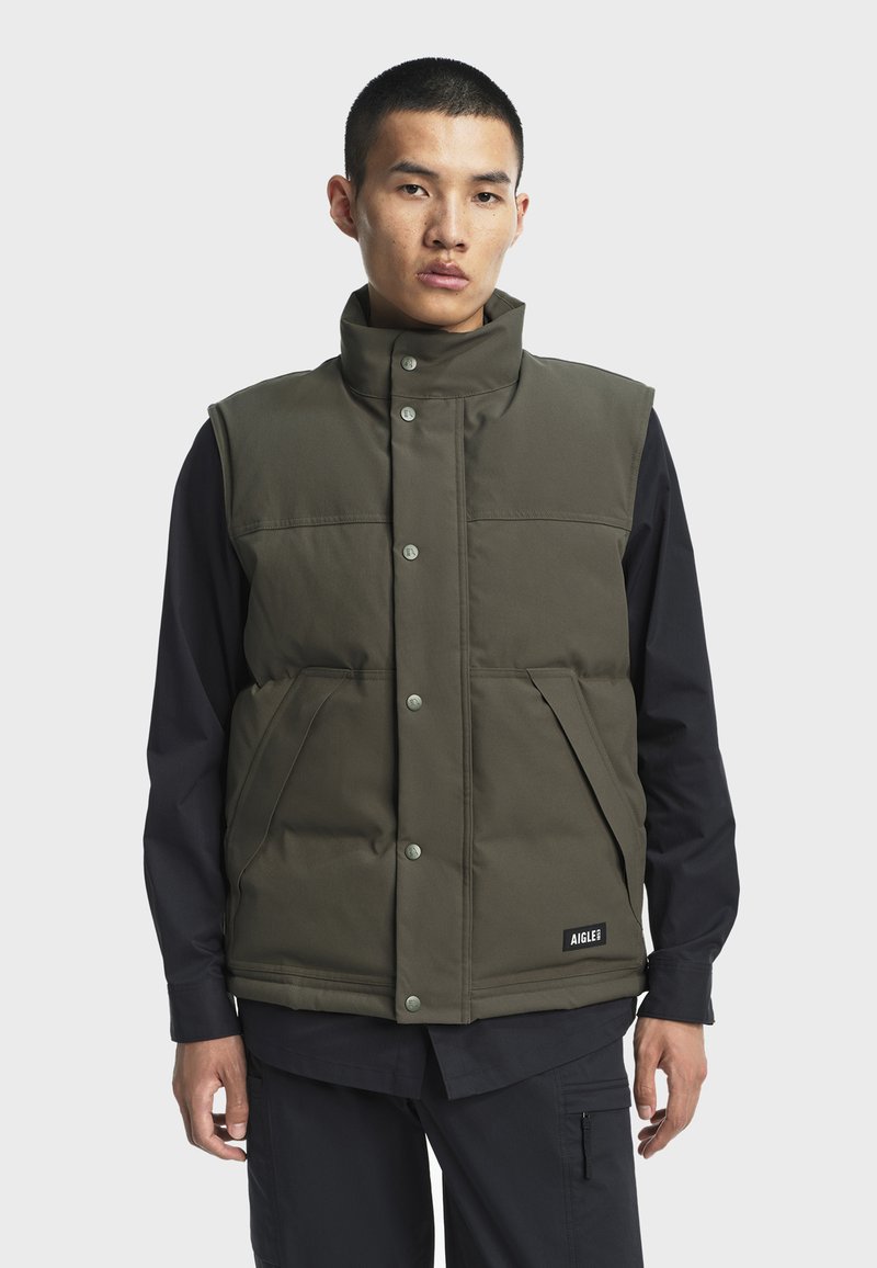 Olivgrüne gepolsterte Weste mit hohem Kragen, Druckknöpfen, zwei Fronttaschen und einem Logo-Label am Saum, getragen über einem schwarzen Langarmshirt.