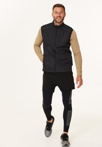 Musta, hihaton liivi korkealla kauluksella, jonka päällä on vaaleanruskea pitkähihaisen paidan, yhdistetty mustiin shortseihin ja mustiin leggingseihin, joissa on harmaita yksityiskohtia.