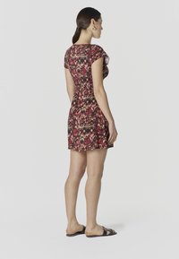 Femme debout, portant une robe à manches courtes rouge et noire à motifs, avec des sandales noires, vue de profil sur un fond uni.