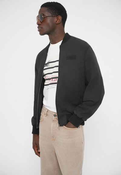 Μαύρο μπουφάν bomber με ριμπ μανσέτες, φερμουάρ μπροστά και λογότυπο στο στήθος, φορεμένο πάνω από άσπρο ριγέ T-shirt και ανοιχτόχρωμα τζιν.
