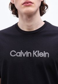 Νέο άτομο με μαλλιά μεσαίου μήκους που φοράει μαύρο μπλουζάκι με το λογότυπο "Calvin Klein" σε λευκό κείμενο στο στήθος.