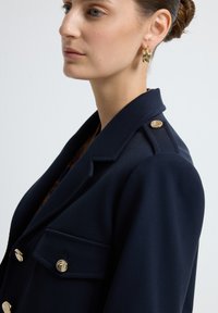 Marineblazer met een gestructureerde snit, voorzien van gouden knopen en een zak met flap. De stof heeft een gestructureerde afwerking, zichtbaar in de kraag en revers.