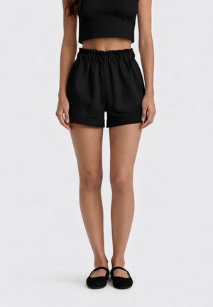 Donna che indossa pantaloncini neri paperbag a vita alta, top nero corto e ballerine nere, in piedi davanti a uno sfondo semplice.