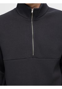 Mörkgrå tröja med hög krage och en silverfärgad dragkedja. Har en texturerad tyg och en enda horisontell söm över bröstet.