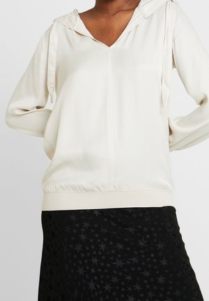 Persoon draagt een crèmekleurige lange mouwen V-hals blouse met een zoomband en een zwarte rok met sterrenpatroon.