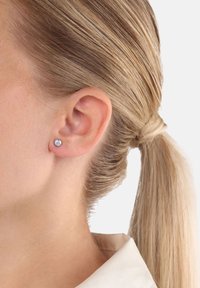 Donna con i capelli biondi legati indietro, che indossa un piccolo orecchino a pavé di diamanti sulla sinistra, su uno sfondo bianco.