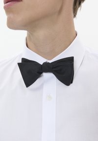 Schwarze Fliege aus strukturiertem Stoff, gelegen auf einem weißen Hemd mit klassischem Kragen und einer einzigen Knopfschließe.