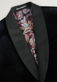 Sort fløjl blazer med en satin sjalkrave og et rigt mønstret bordeaux inderside. Teksturen står i kontrast til det glatte stof.
