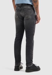 Zwarte spijkerbroek met een tapering pasvorm, voorzien van vervaagde details, twee achterzakken en een zichtbare merketiket op de taille. Draagt met zwarte laarzen.