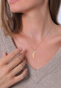 Goldene Halskette mit einem Federanhänger und einem kleinen Strasssteinbesatz, an einer dünnen Kette hängend, getragen über einem beigen Pullover.
