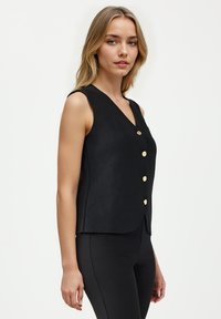 Czarna bezrękawnik z dekoltem w kształcie litery V i złotymi guzikami. Wykonana z teksturowanej tkaniny, z dopasowanym krojem i lekko zaokrąglonym dołem.