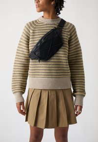 Black nylon crossbody bag with a curved design, front zipper, and adjustable strap, worn over a striped sweater and pleated skirt.