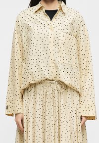 Chemise beige à boutons et jupe à pois noirs portée sur un top noir, vue de face des épaules à la taille.