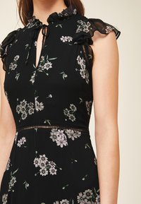 Svart blommig klänning med volangkrage, blommönster i ljusa färger och genomskinliga volangdetaljer på axlarna, med en nyckelhålsöppning framtill.