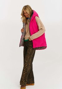 Jasně růžová nadýchaná vesta s leopardím vzorem uvnitř, nošena přes béžový svetr a zelené akcenty, kombinována s flared kalhotami s leopardím vzorem.