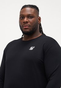Black long-sleeve shirt made of soft cotton, featuring a white embroidered logo on the left chest, round neckline, and relaxed fit.