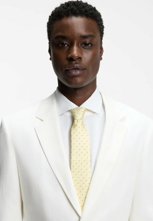 Mand med kort krøllet hår iført en hvid dragt, hvid skjorte og en bleg gul slips med små sorte prikker, set forfra.