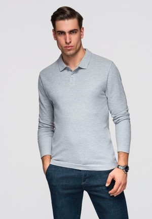 Mann mit zurückgekämmtem Haar, trägt ein hellgraues, langärmliges Poloshirt und dunkelblaue Jeans, posiert mit einer Hand in der Tasche und trägt eine Armbanduhr.