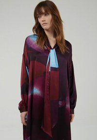 Langermige jurk met een diep bordeauxrode basis, afgewerkt met abstracte blauwe en rode patronen, satijnen textuur en een zelfbindende kraagdetail.