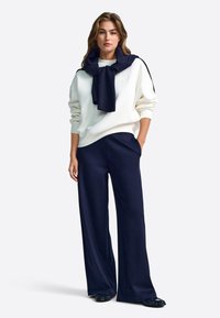 Balta lielizmēra sporta krekls ar tumši zilu šalli ap kaklu, kombinēts ar tumši ziliem vaļīgām biksēm un spīdīgām melnām kurpēm.