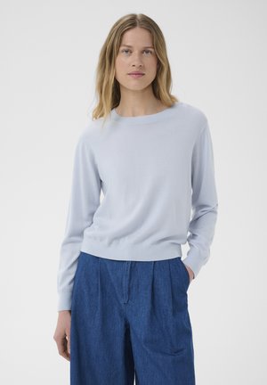 Blonde vrouw in een lichtblauwe trui met lange mouwen en een losse blauwe broek met hoge taille, met één hand in de zak.
