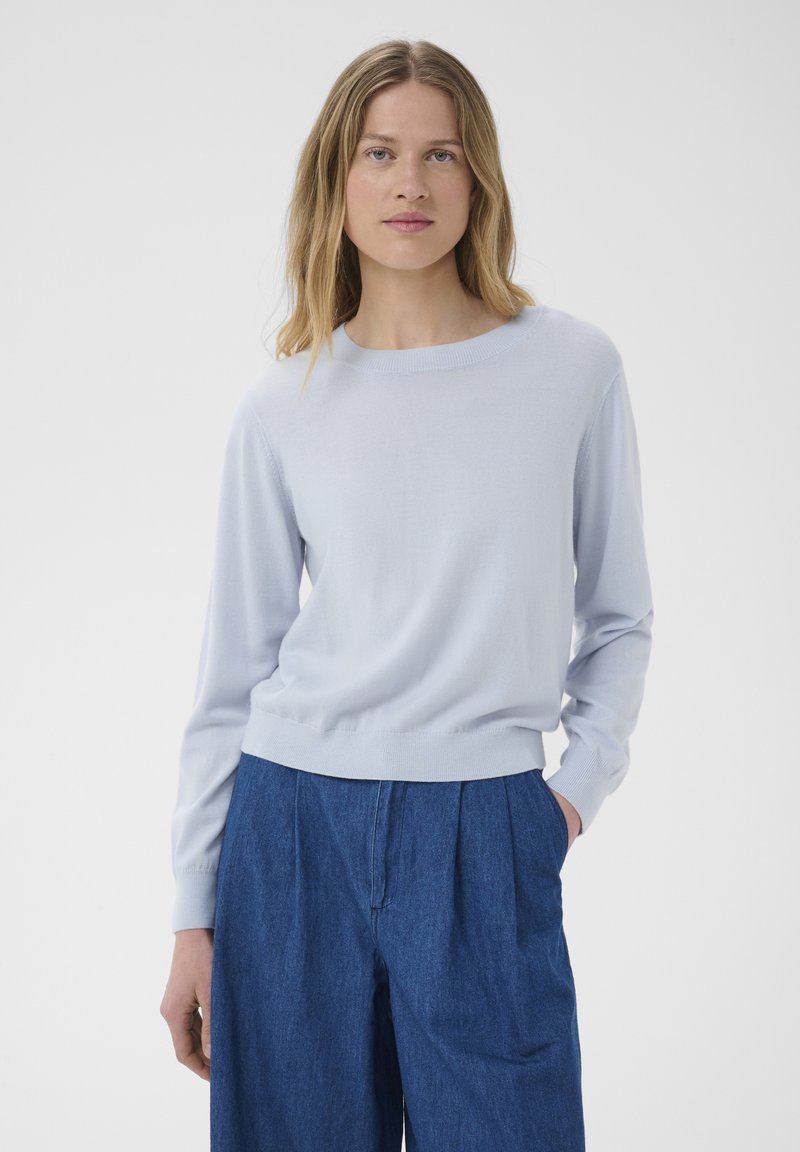 Femme blonde portant un pull bleu clair à manches longues et un pantalon bleu ample taille haute avec une main dans la poche.