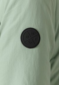 Patch circulaire noire avec les lettres N, M, T et le chiffre 97 séparés par des lignes, sur un fond de tissu vert clair.