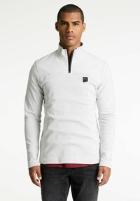 Hvid, langærmet pullover med en kvart lynlås og et sort logo-patch på brystet. Har en glat tekstur og en tætsiddende design.