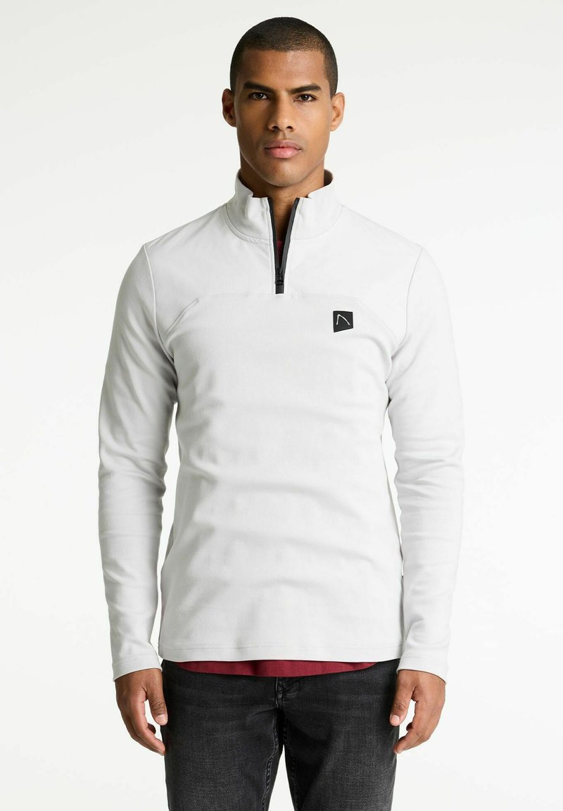 Pullover bianco a maniche lunghe con zip a un quarto e un patch nero sul petto. Presenta una texture liscia e un design aderente.