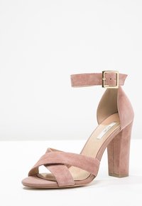 Rosa mockakänga med klack, rem vid vristen, guldfärgad spänne och korsade framskydd på vit bakgrund.