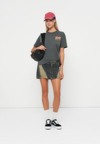 Kobieta w czerwonej czapce, czarnej koszulce AC/DC, patchworkowej mini spódniczce z dżinsu, białych skarpetkach, czarnych sneakersach i niosąca czarną torbę na ramię.
