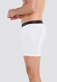 Weiße Boxershorts aus weichem Stoff, mit einem schwarzen elastischen Bund und eingeprägtem Logo. Seitenansicht, die Passform und Design zeigt.
