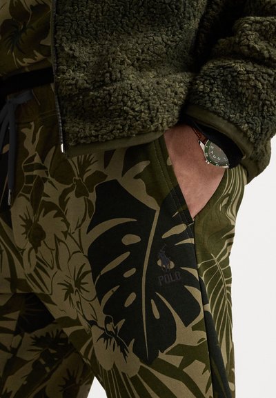 Chaqueta de forro polar verde oliva con un acabado texturizado, combinada con pantalones verde oliva oscuros que presentan un patrón de hojas y un logotipo discreto.