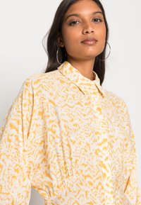Camisa com padrão amarelo e branco, com gola e um corte solto, apresentando uma textura subtil e um design de ombros caídos.