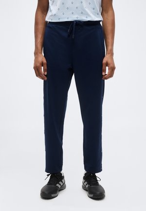 Personne portant un pantalon à cordon de serrage bleu marine et des chaussures de sport noires avec des bandes blanches, debout devant un fond uni.