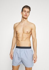 Pruhované modré boxerky s černým elastickým pasem, s volným střihem a měkkou texturou látky, předváděné osobou bez trička.