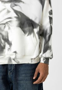 Witte en grijze tie-dye hoodie met een verstelbare kap, steekzak en geribde manchetten. Gepaard met donkerblauwe jeans.