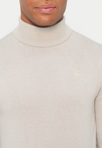 Beżowy sweter typu golf wykonan z prążkowanej dzianiny, charakteryzujący się dopasowanym krojem i małym, tonalnym haftowanym logo na piersi.