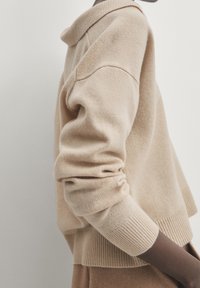 Pull beige à manches longues avec détails aux poignets porté par une personne sur un fond clair uni.