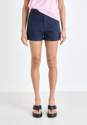Personne portant un short bleu marine à revers, une chemise rayée rose et blanche, et des sandales compensées noires, debout devant un fond blanc uni.