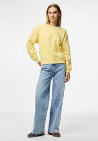 Felpa di cotone gialla con collo a giro, spalle scese e polsini a costine, abbinata a jeans a gamba larga in denim chiaro e scarpe marroni.
