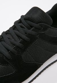 Baskets noires de sport présentant un tissu texturé et du daim lisse, avec des lacets plats et une semelle en caoutchouc blanche. Design à bout rond.