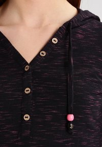 Dunkelgrauer Kapuzenpullover mit Holzknöpfen vorne und einem Kordelzug, der nahe dem Ausschnitt mit pinken und schwarzen Perlen verziert ist.