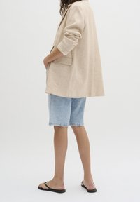 Beżowa lniana marynarka z usztywnionymi ramionami, pojedynczą klapą kieszeniową, zestawiona z jasnoniebieskimi denimowymi szortami i czarnymi japonkami.