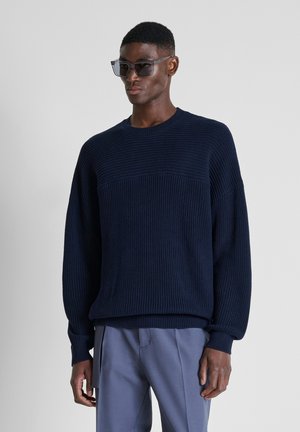 Man die een donkerblauwe gebreide trui, lichtblauwe broek en donkere zonnebril draagt, staat tegen een effen lichtgrijze achtergrond.