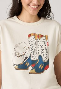 T-shirt créme à manches courtes présentant un graphisme de bottes de cowboy bleues avec des accents dorés et un chapeau blanc avec un bandeau coloré.